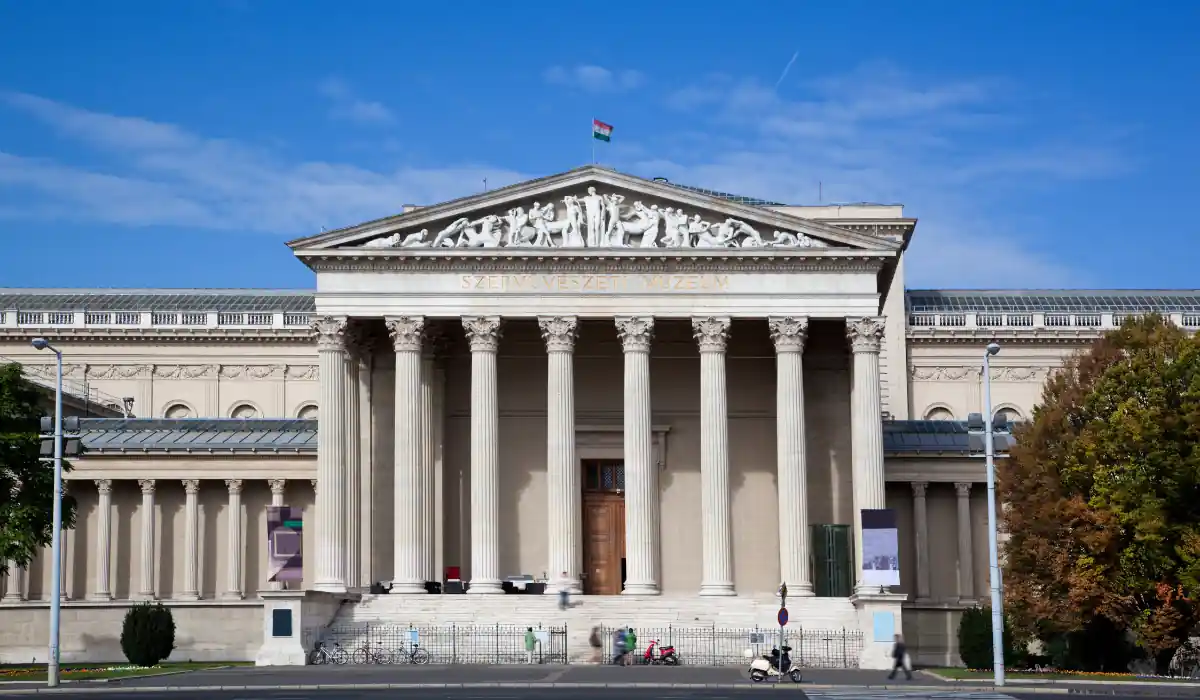 The 15 Best Things to Do in New Orleans for Couples 18 The image shows the front facade of the Museum of Fine Arts in Budapest. It features large Corinthian columns, detailed sculptures on the pediment, and a flag flying on top. A clear blue sky is in the background.