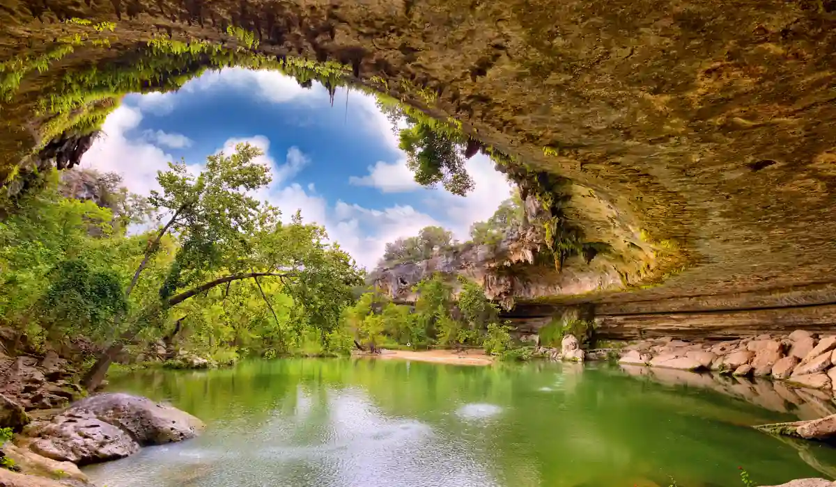 Top 15 Fun $ Unique Places That Don't Feel Real in the USA 2025 17 A serene natural pool beneath a rocky overhang, surrounded by lush greenery and trees. Sunlight filters through, reflecting on the calm green water. The sky is partly cloudy, adding depth to the vibrant scene.