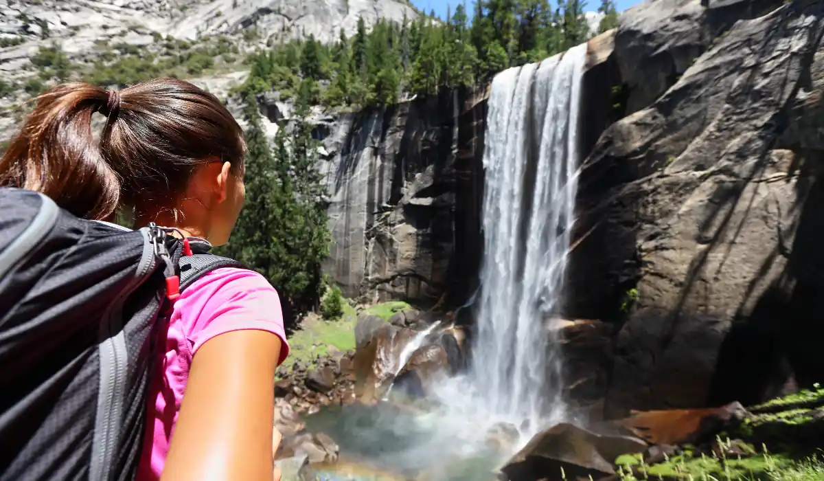 20 Most Romantic Things to do in California for Couples 9 A person wearing a pink shirt and backpack stands in the foreground, gazing at a waterfall cascading down rocky cliffs surrounded by forested mountains on a sunny day.