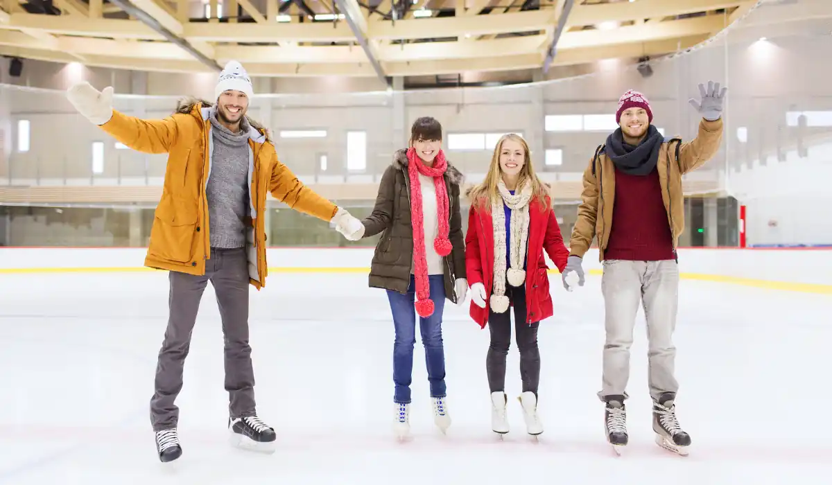 40 amazing Cute Date Ideas in Columbus, Ohio for Couples in 2025 23 Four people holding hands and smiling while ice skating indoors. They are dressed warmly in winter clothes, including jackets, hats, and scarves. The ice rink has wooden beams and large windows in the background.
