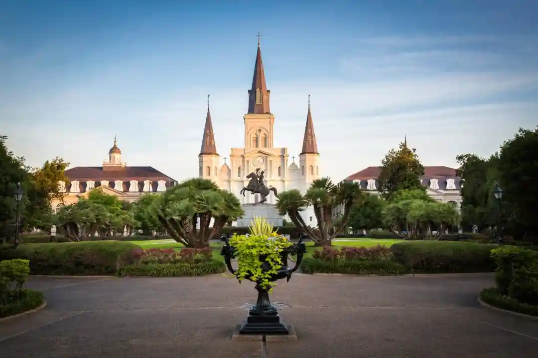 View of Jackson Square in New Orleans with a prominent equestrian statue surrounded by greenery. St. Louis Cathedral stands majestically in the background under a clear blue sky.
