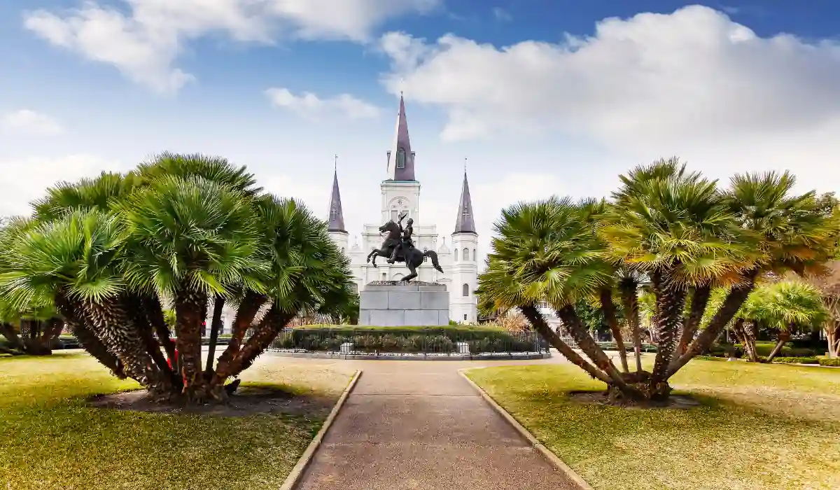 The 15 Best Things to Do in New Orleans for Couples 5 A central path flanked by palm trees leads to an equestrian statue with a historic cathedral in the background. The sky is partly cloudy, adding a bright ambiance to the scene.