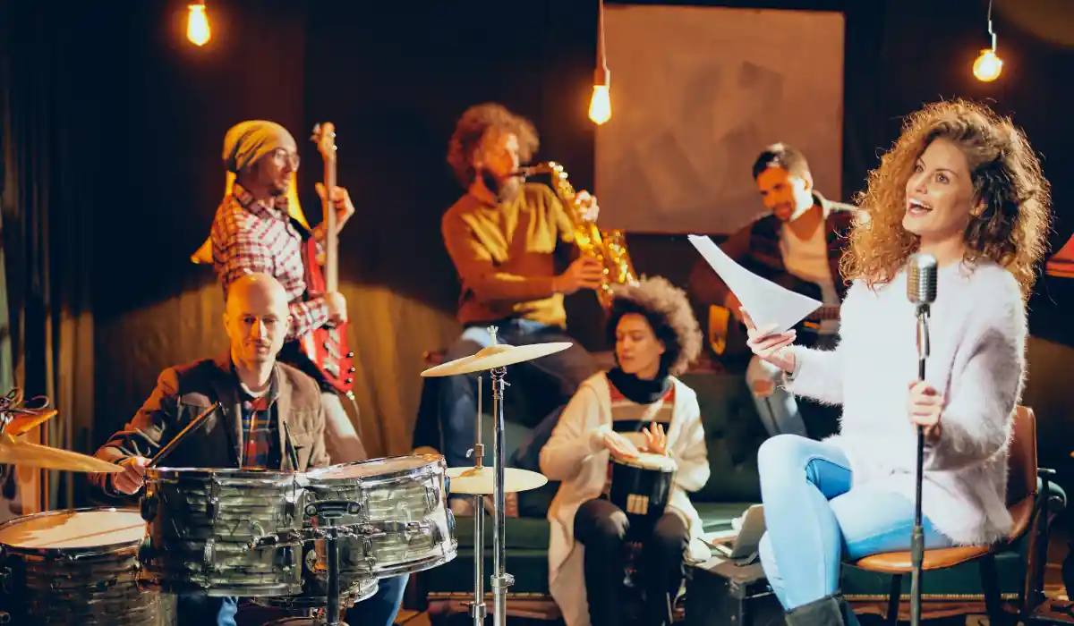The 15 Best Things to Do in New Orleans for Couples 13 A band is performing in a cozy, dimly lit space. A drummer and a singer with sheet music are in the foreground. A guitarist, saxophonist, and two other musicians play in the background, creating an intimate and lively atmosphere.