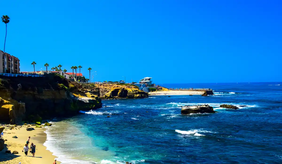 12+ Best Things to do in San Diego for Couples 2025 3 Scenic view of a rocky coastline with clear blue water and a sandy beach. People are enjoying the sun and ocean, while a few palm trees and buildings line the shore. The sky is a vibrant blue, complementing the tranquil sea.
