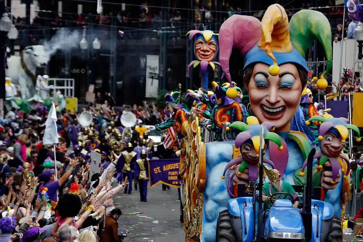A colorful parade float with a jester theme travels down a street during a Mardi Gras celebration. The float features large jester heads, and people in costumes wave from it. A crowd of onlookers lines the street, reaching out with excitement.
