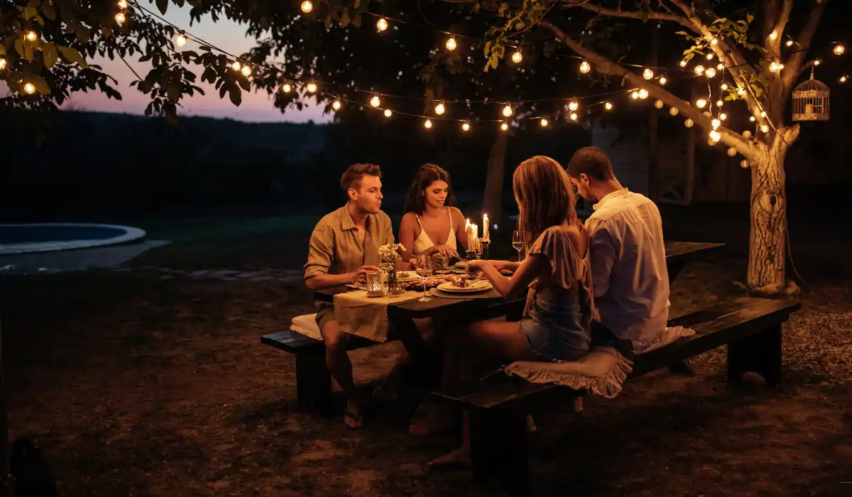 40 amazing Cute Date Ideas in Columbus, Ohio for Couples in 2025 6 Four people sit at a wooden picnic table enjoying a candlelit dinner outdoors under string lights. The scene is set during twilight, creating a warm, inviting atmosphere. Trees and soft lighting add to the cozy ambiance.