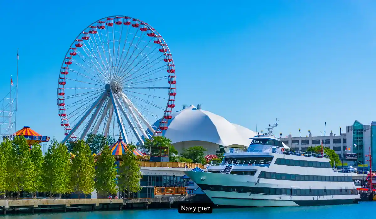 Top 15 Romantic Things to Do in Chicago for Couples 13 A sunny view of Navy Pier featuring a large Ferris wheel, a white domed building, and trees lining the waterfront. A white and blue passenger boat is docked by the pier.
