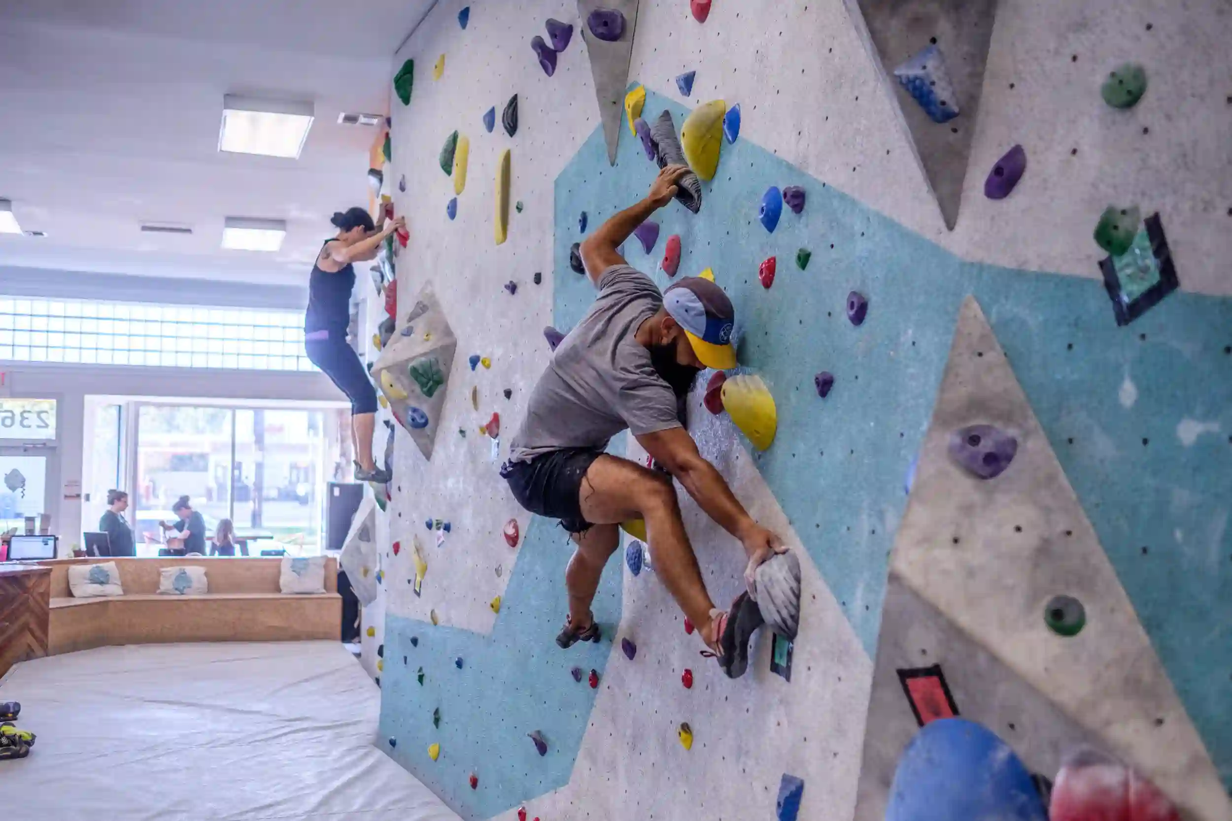 Two people are bouldering on an indoor climbing wall with colorful holds. The foreground climber wears a cap and gray shirt, focusing on a dynamic movement. A person in the background appears to be reaching for a hold. Bright interior lighting.
