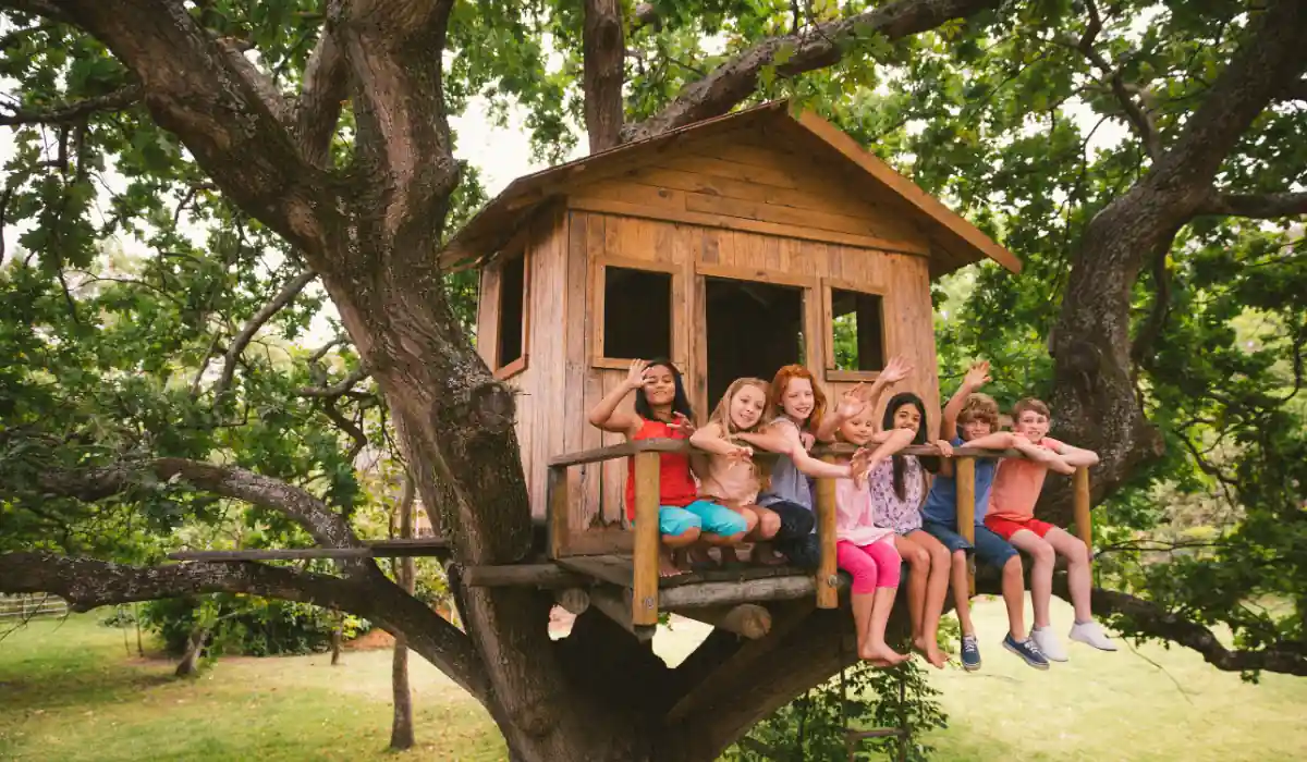 20 Most Romantic Things to do in California for Couples 19 A group of children sit on the railing of a wooden treehouse built in a large tree. They smile and wave at the camera, surrounded by lush green leaves. The treehouse has small windows and a simple design, conveying a fun, adventurous atmosphere.