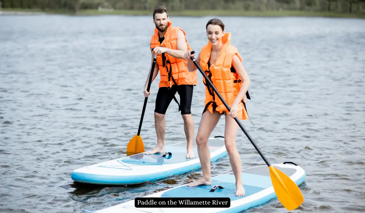 Top 15+ Romantic Things to do in Portland for Couples 4 Two people wearing orange life vests are stand-up paddle boarding on a calm river. They each hold a paddle, and the water stretches out behind them with trees lining the distant shore. A caption reads, "Paddle on the Willamette River.