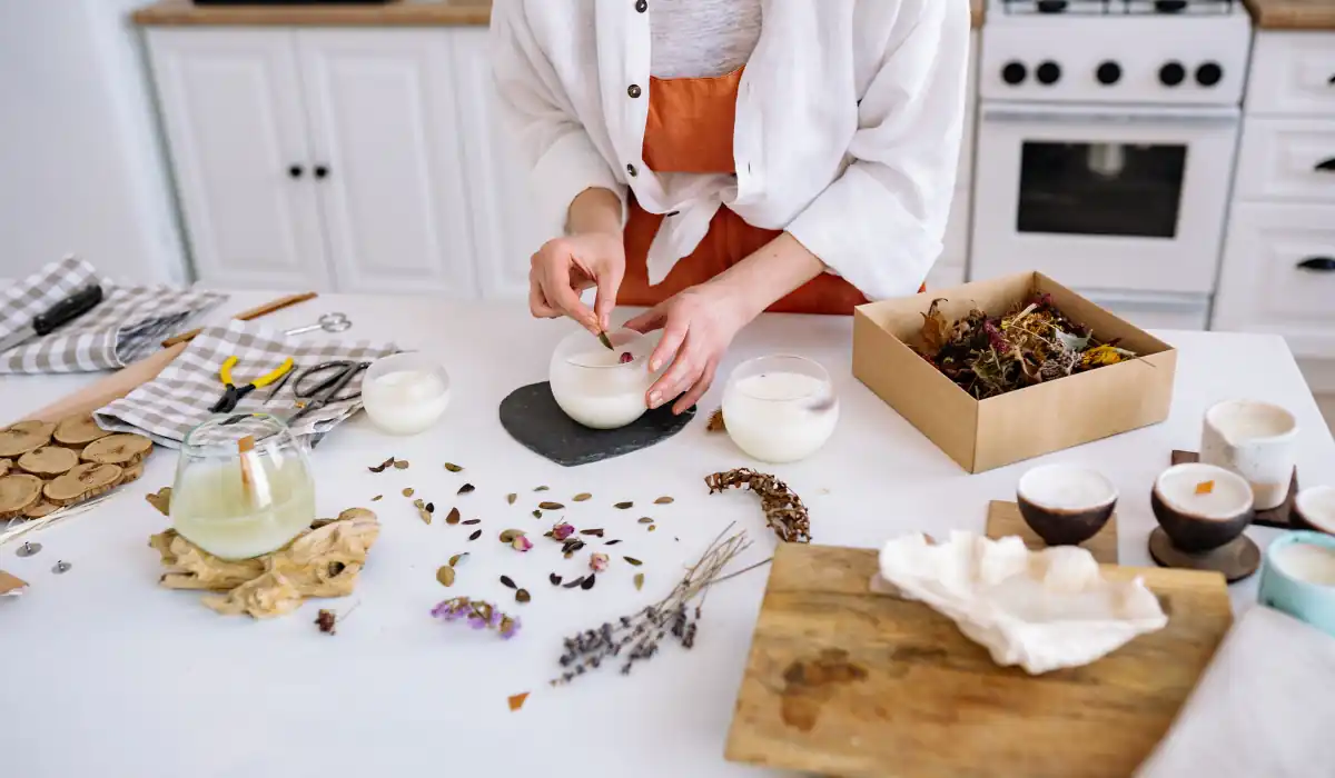 40 amazing Cute Date Ideas in Columbus, Ohio for Couples in 2025 8 A person stands in a kitchen pouring wax into glass containers to make candles. The countertop is covered with dried flowers, herbs, tools, and a box of additional materials. The background shows white cabinets and a stove.