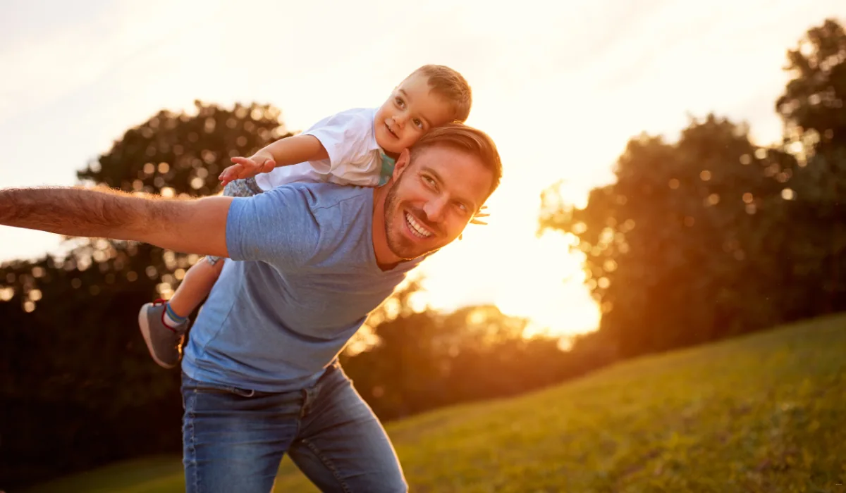 40 amazing Cute Date Ideas in Columbus, Ohio for Couples in 2025 33 A smiling man in a blue t-shirt gives a little boy a piggyback ride in a sunlit park. The boy stretches his arms out, both appearing joyful against the backdrop of trees and a setting sun.