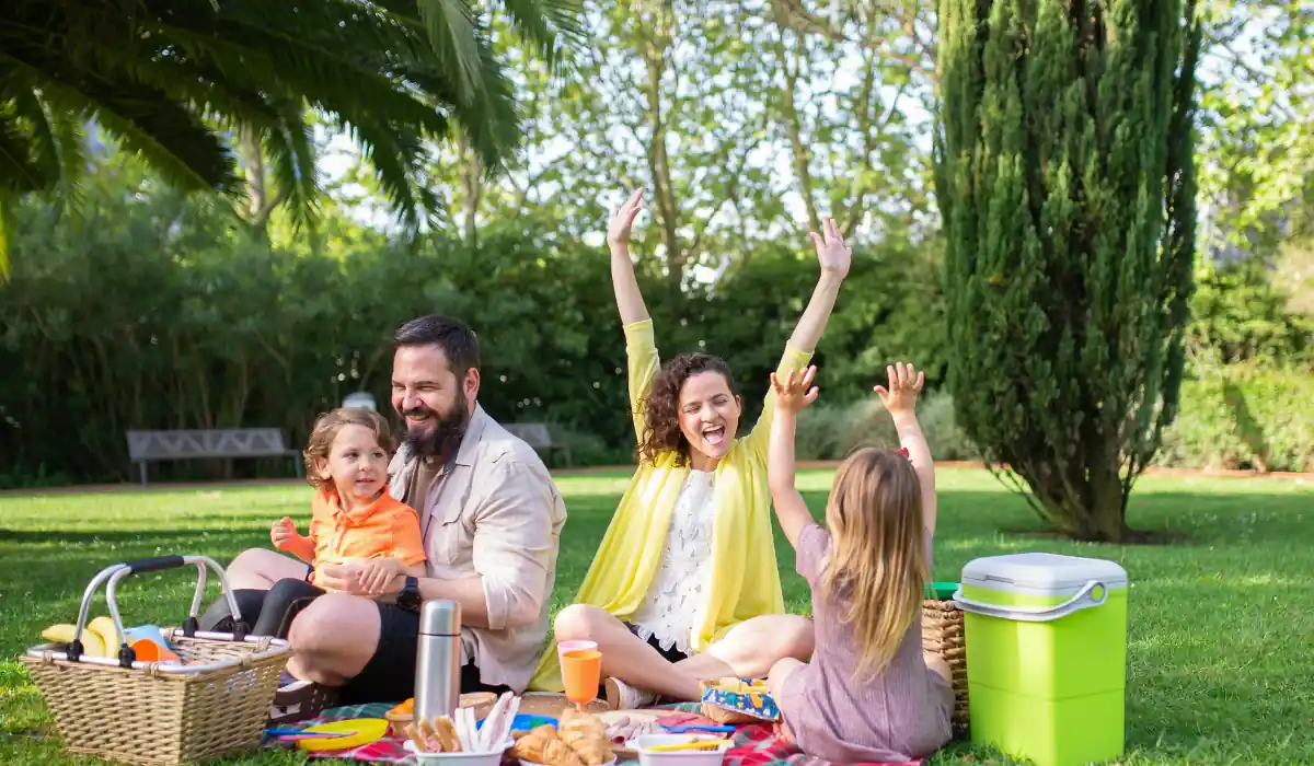 15 Free Romantic Things to Do in Massachusetts for Couples 10 A family enjoys a picnic on a sunny day in a park. The parents and two children sit on a blanket surrounded by food and drinks. The mother and daughter raise their arms in joy, with trees and greenery in the background.