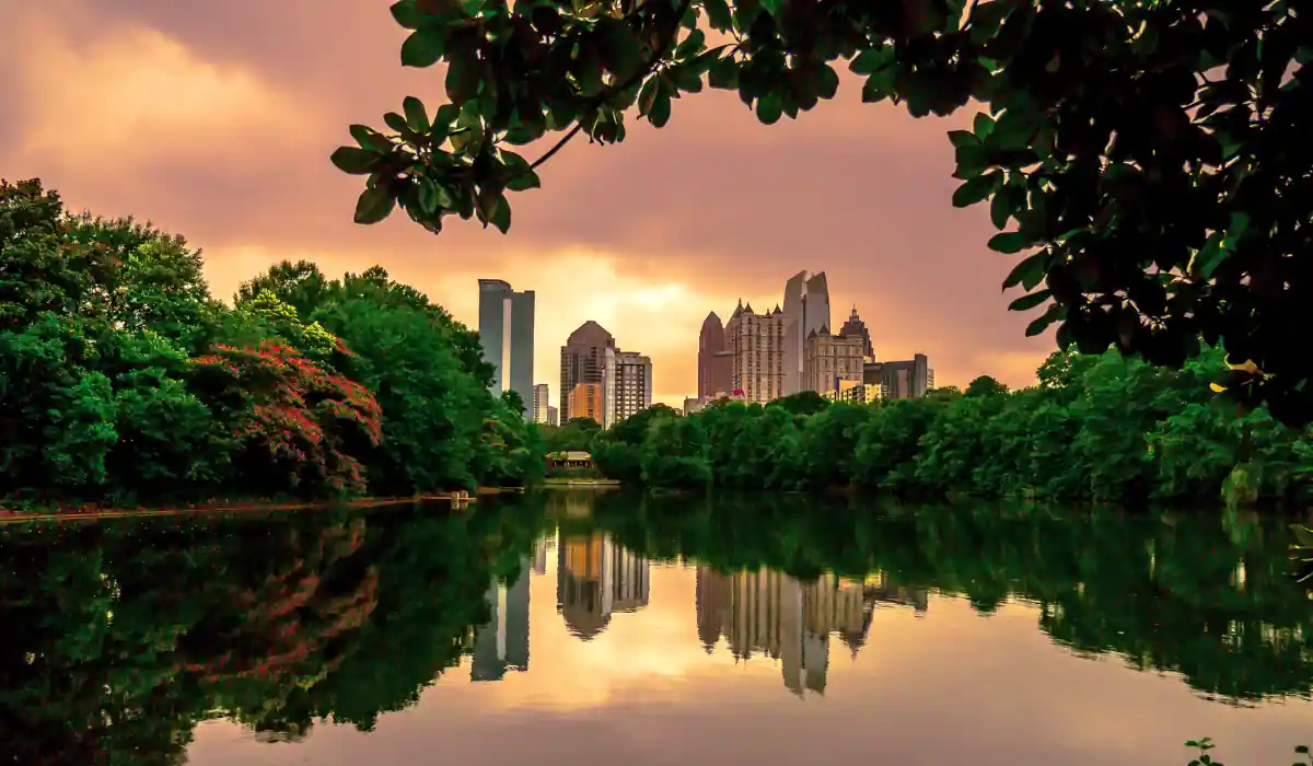 Top 15+ Free Things to do in Atlanta for Couples 2025 4 A city skyline is reflected in a calm lake surrounded by lush greenery. The sky is an orange hue, suggesting a sunset or sunrise. The scene is framed by leafy branches at the top, creating a tranquil urban nature landscape.