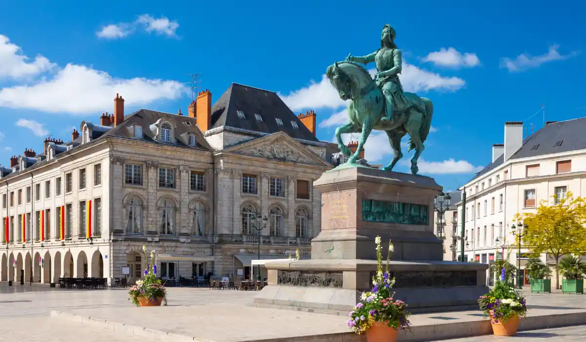 A historic town square featuring a large equestrian statue in the center. Surrounding the statue are elegant, classic-style buildings with steep roofs and ornate facades. Bright flowers decorate planters nearby under a clear blue sky with a few clouds.
