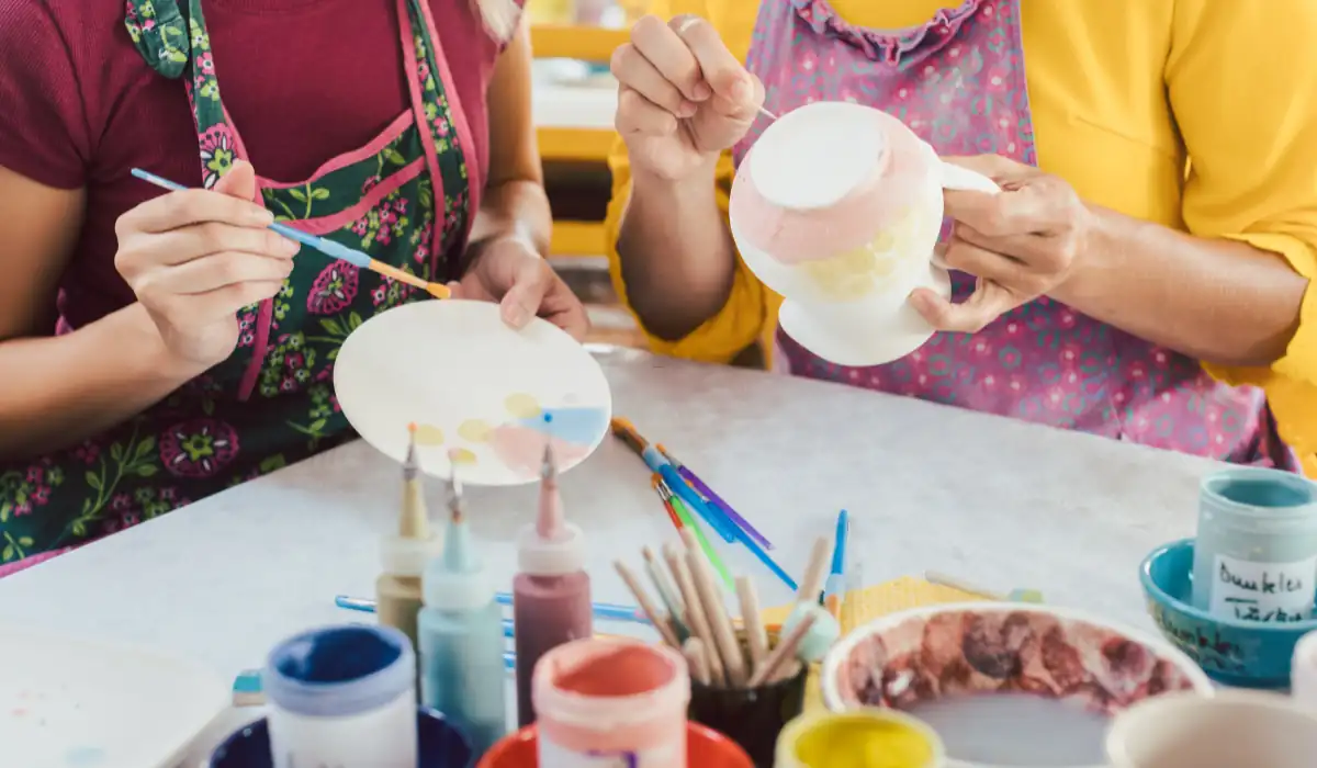 40 amazing Cute Date Ideas in Columbus, Ohio for Couples in 2025 10 Two people painting ceramic items at a table. One holds a round plate, and the other holds a cup. Various paints and brushes are on the table. They are wearing aprons, and the atmosphere is colorful and creative.