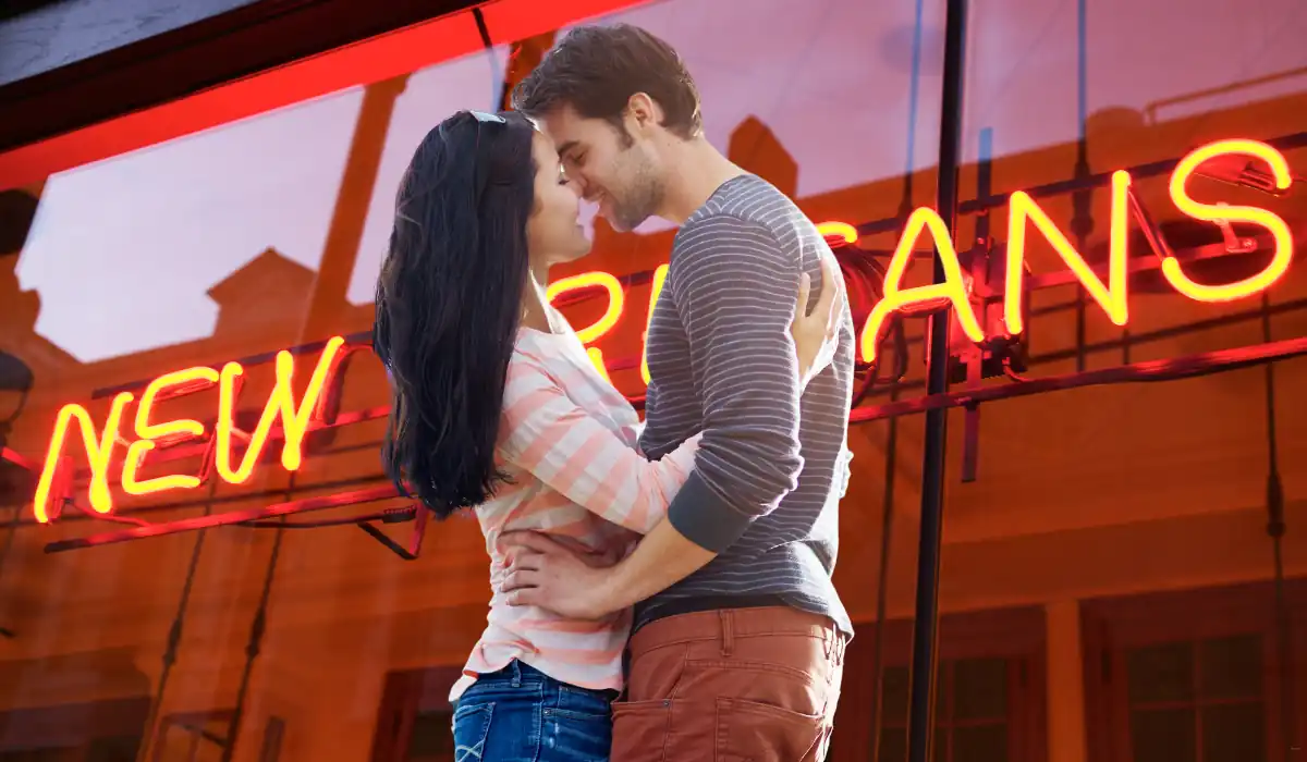 The 15 Best Things to Do in New Orleans for Couples 17 A couple embraces lovingly in front of a red neon sign that reads NEW ORLEANS. They smile at each other, enjoying a moment together. The signs glow reflects on the glass behind them. The setting suggests a lively, urban atmosphere.