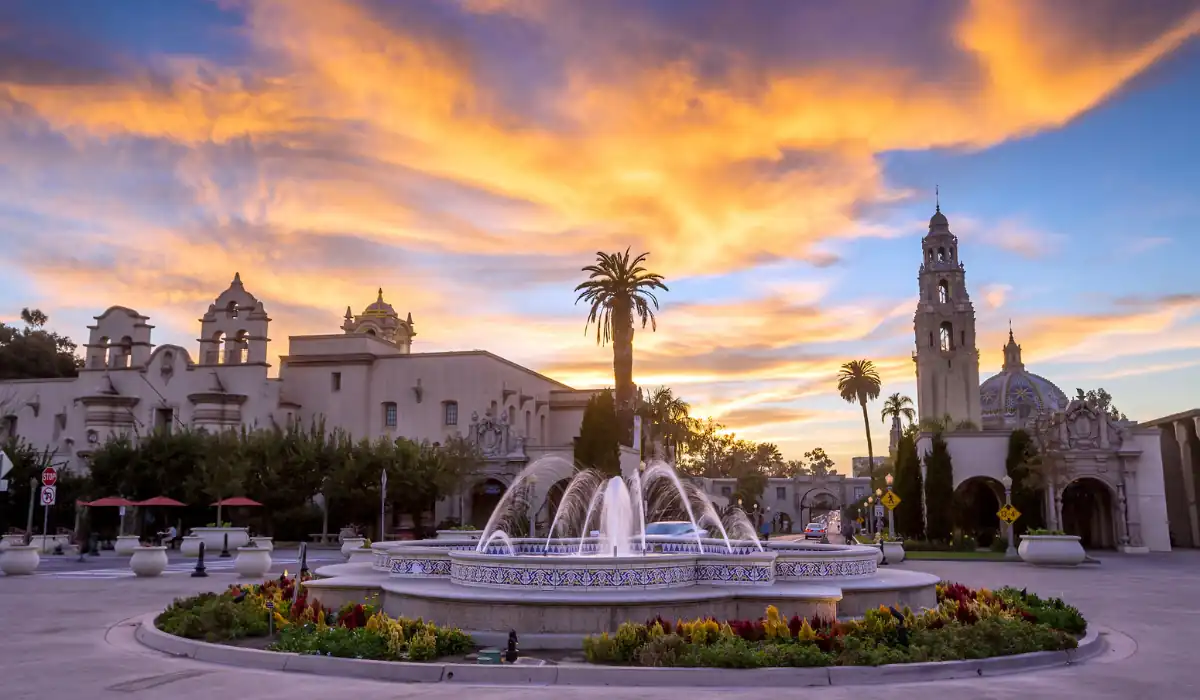 A vibrant sunset sky with orange clouds over a historic building and a fountain in a landscaped area. Tall palm trees and flowering plants are in the foreground. "Romantic Things to do in San Diego for Couples"