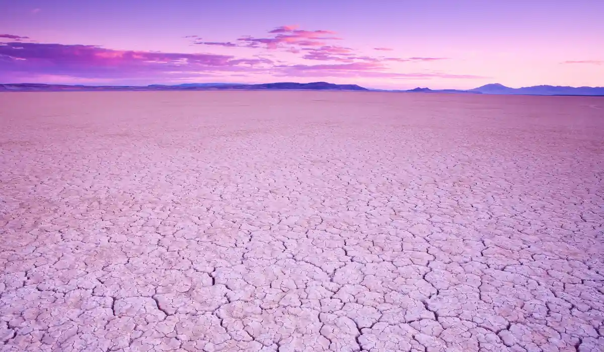 Top 15 Fun $ Unique Places That Don't Feel Real in the USA 2025 9 A vast, cracked desert landscape under a vibrant purple and pink sky at sunset. Mountains are visible on the horizon, creating a stark contrast with the desolate, textured earth in the foreground.