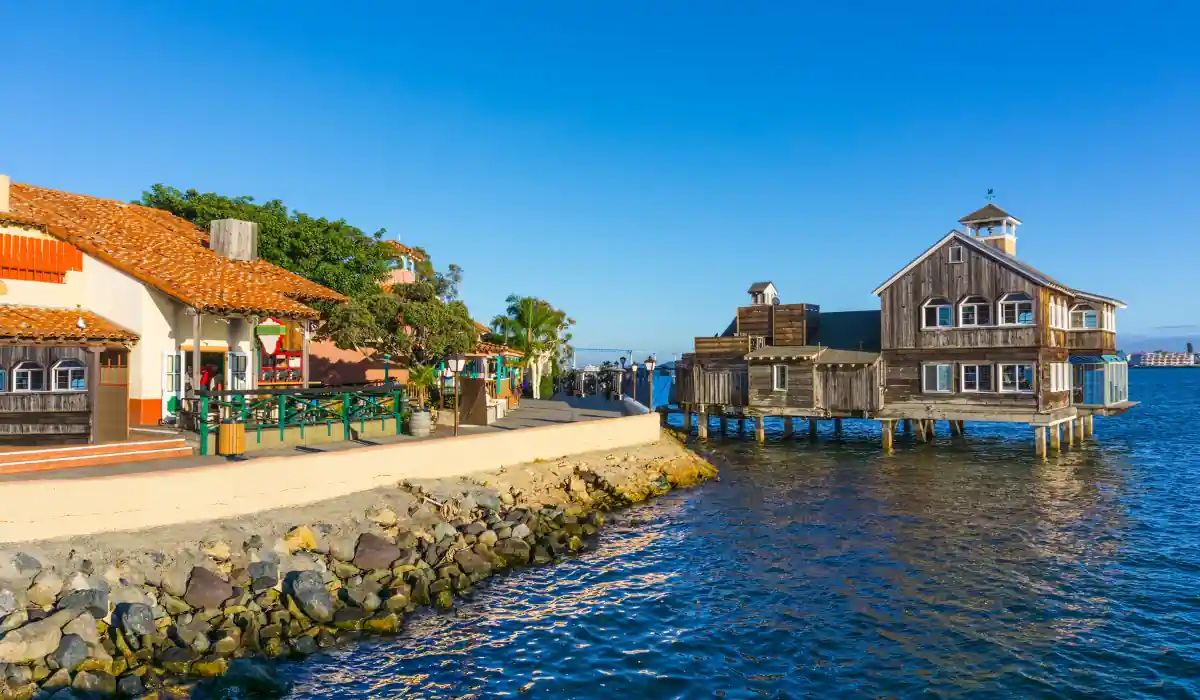 12+ Best Things to do in San Diego for Couples 2025 8 A picturesque seaside village with rustic buildings, one on stilts over the water. The foreground features a rocky shoreline under a clear blue sky, enhancing the tranquil coastal ambiance.