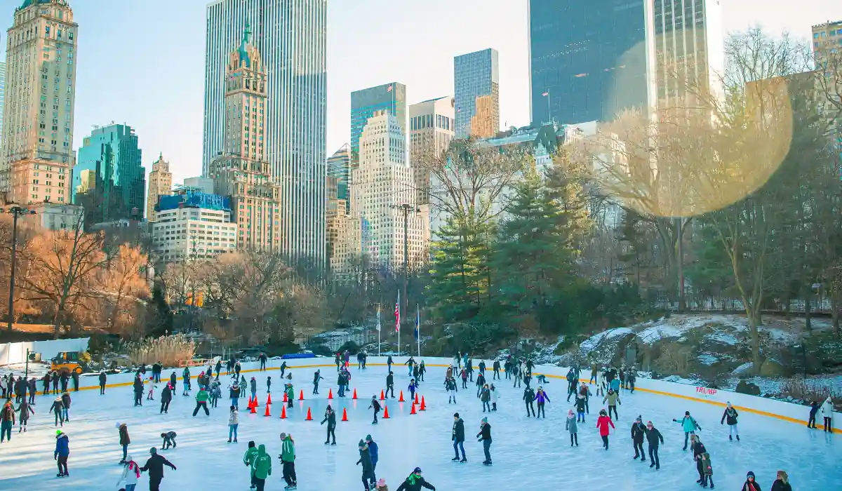 Top 15 Romantic Things to Do in Chicago for Couples 5 People ice skating on a large outdoor rink in a park, surrounded by skyscrapers and trees. Cones organize skaters, and sunlight shines through, creating a bright winter scene.