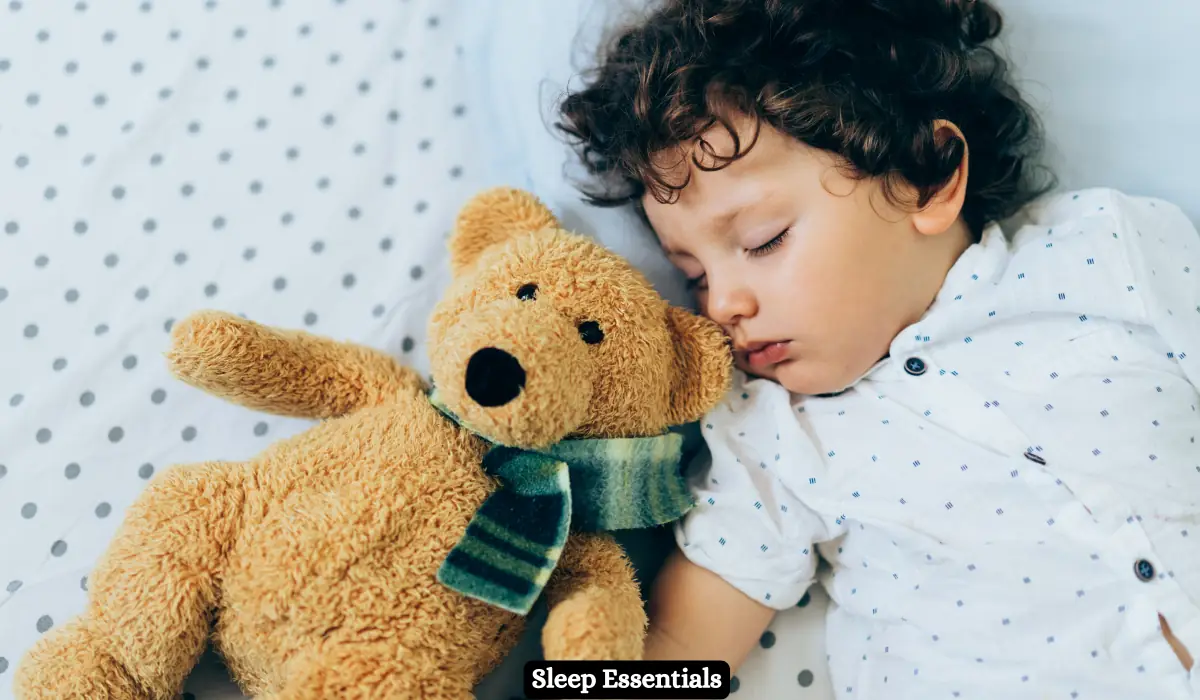 Toddler Travel Essentials for Road Trip in Winter 2025 8 A young child with curly hair sleeps peacefully on a polka dot bed sheet, cuddling a brown teddy bear wearing a striped scarf. The child is wearing a white shirt with small black polka dots. At the bottom, there is text that reads "Sleep Essentials.