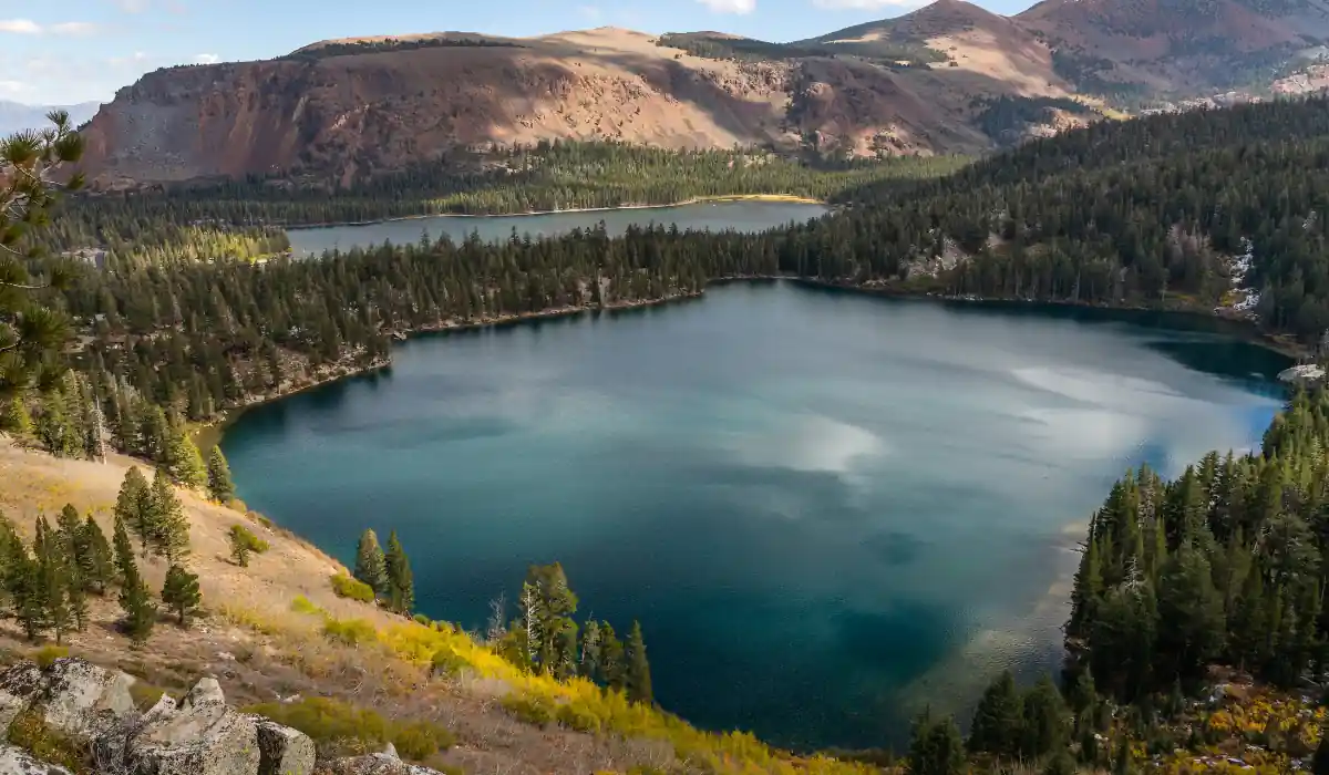 20 Most Romantic Things to do in California for Couples 13 A serene alpine lake surrounded by dense evergreen forests and rolling hills. The water is a deep blue, reflecting the partly cloudy sky above. In the background, a second lake and rugged mountains rise gently.