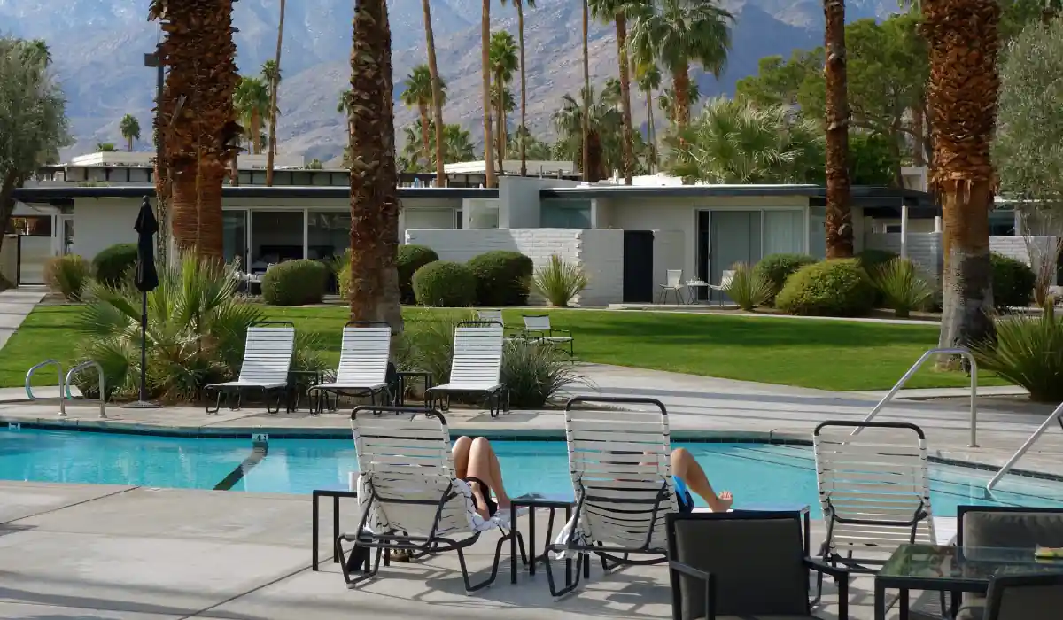 20 Most Romantic Things to do in California for Couples 7 Two people reclining on lounge chairs by a pool, surrounded by tall palm trees and manicured greenery. A modern building with large glass windows is visible in the background, set against a backdrop of mountains.