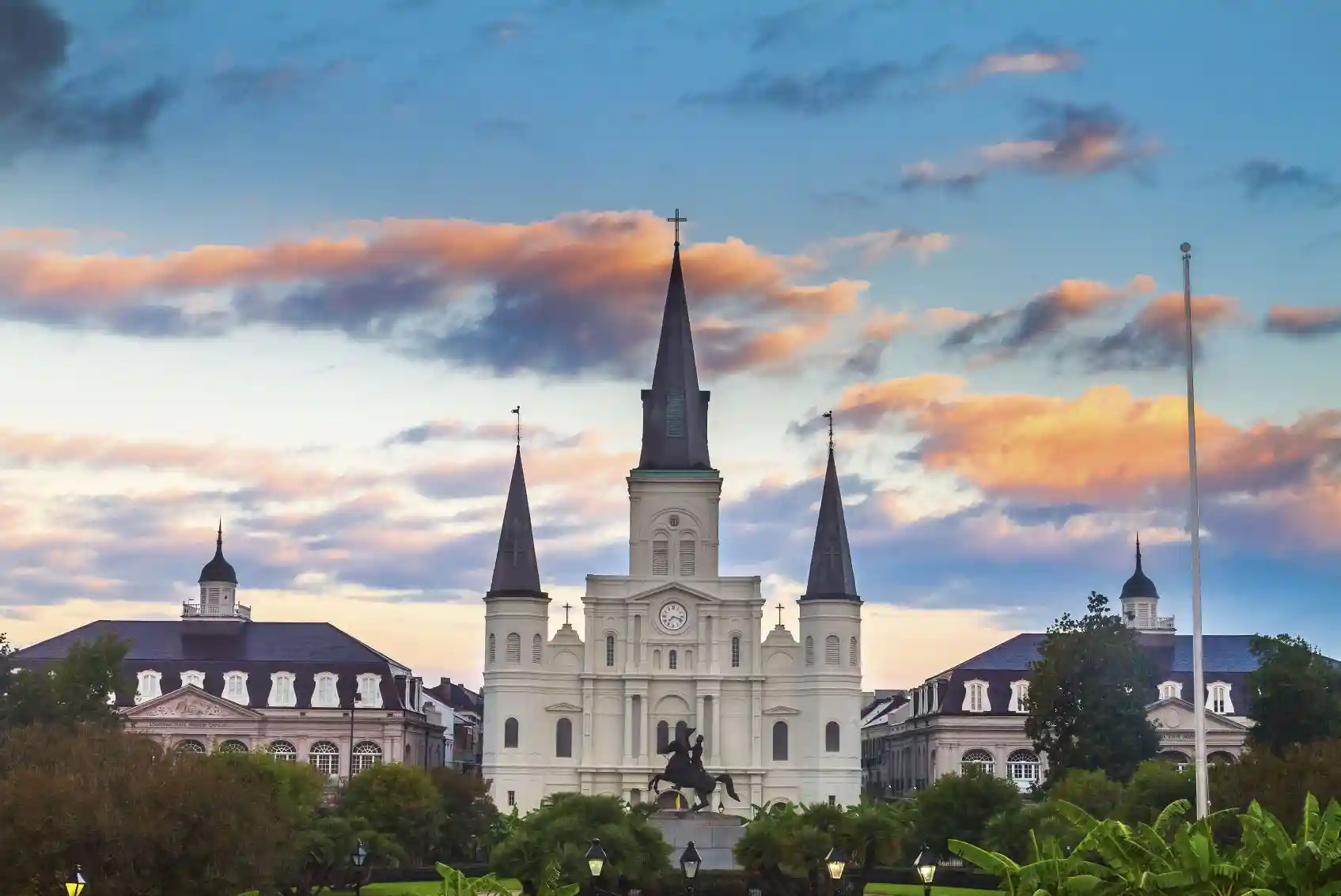 A grand cathedral with multiple spires stands against a vibrant sky at sunset. In the foreground, a statue of a man on horseback is silhouetted by the warm, colorful clouds. Trees and buildings encircle the scene, creating a serene and historical ambiance.
