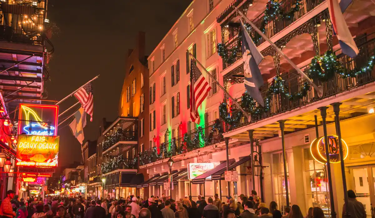 The 15 Best Things to Do in New Orleans for Couples 20 A lively, crowded street at night with buildings adorned in colorful lights and decorations. Flags hang from balconies, and the bright glow of neon signs illuminates the scene, creating a festive atmosphere.