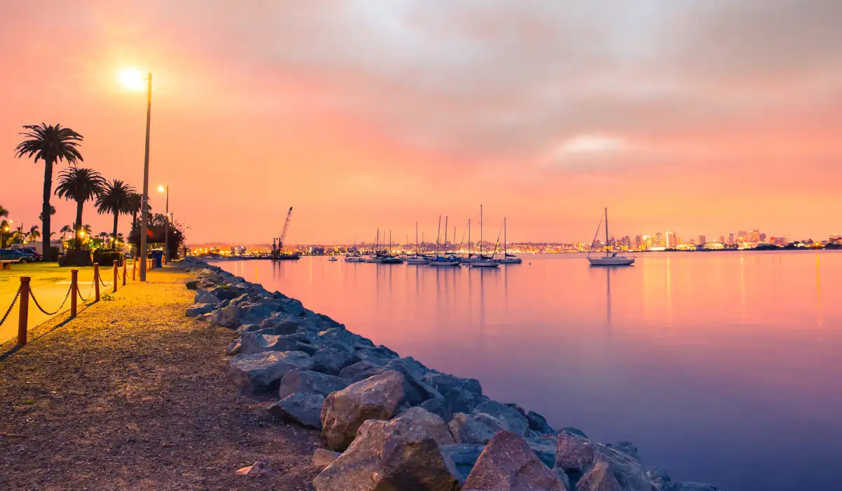 20 Most Romantic Things to do in California for Couples 3 A serene waterfront scene during sunset, with a path lined by palm trees and streetlights on the left and a rocky shore on the right. Boats are docked in the calm water, and the distant city skyline is illuminated under an orange sky.