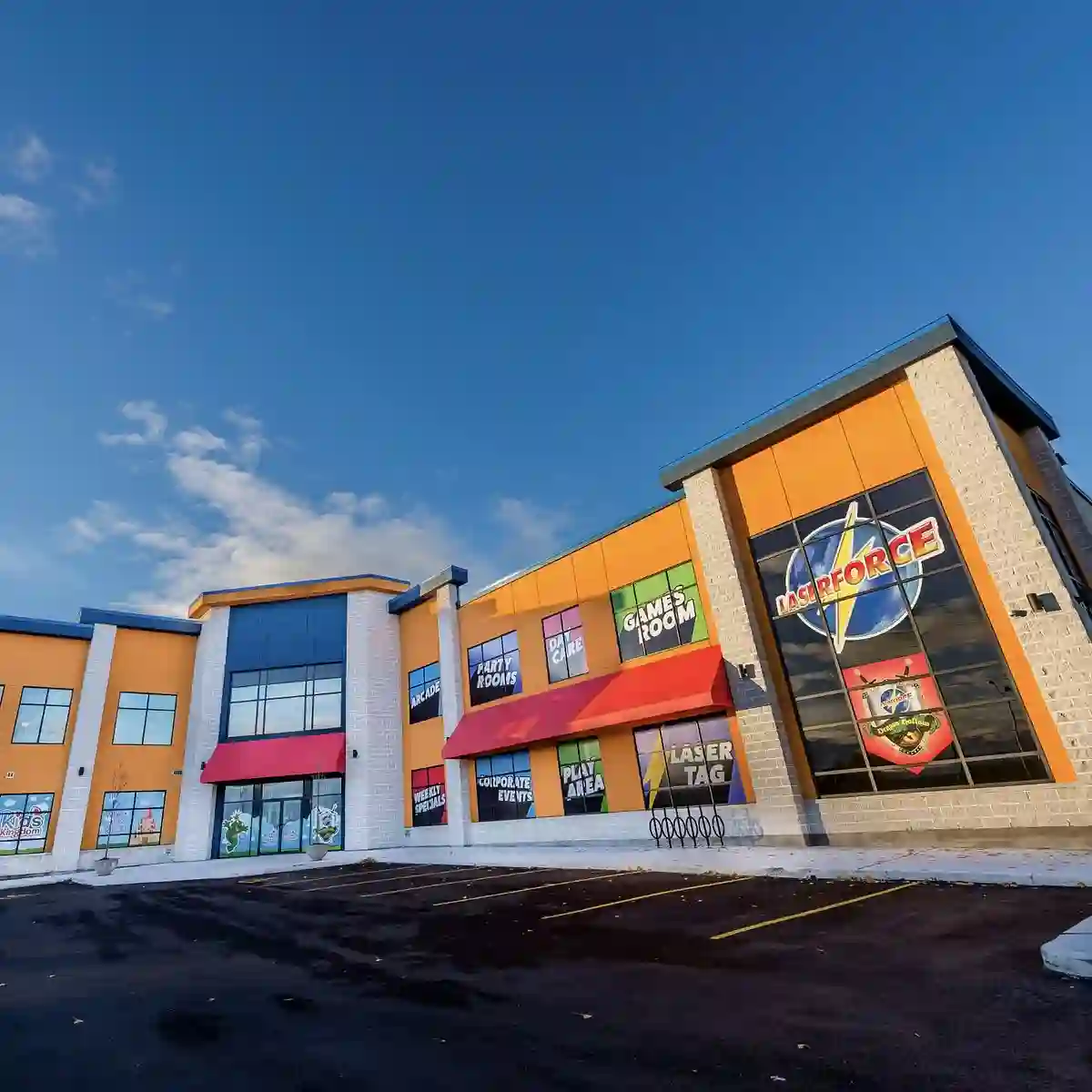 Exterior view of an entertainment center with colorful signage for activities like laser tag and a game room. The building features large windows, red awnings, and a spacious parking lot under a blue sky with scattered clouds.
