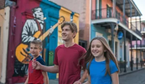Three kids walk arm in arm down a street with colorful murals. One mural features a musician with a guitar. The buildings have balconies and vibrant facades, creating a lively, artistic atmosphere. "Things to Do in Orleans With Kids"