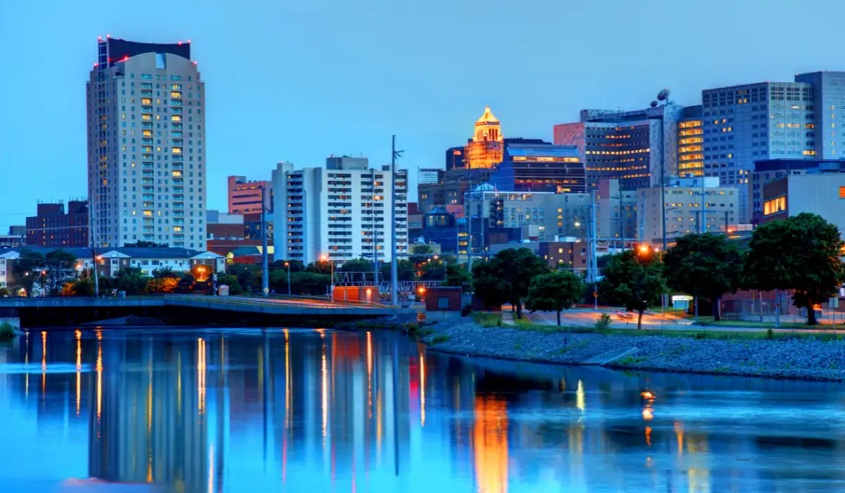 City skyline at dusk featuring tall buildings with lights on. A river reflects the illuminated structures and a bridge crosses the water in the foreground. Trees line the riverbank, and the sky is a soft blue as evening settles in. "Things to Do in Rochester for Couples at Night"
