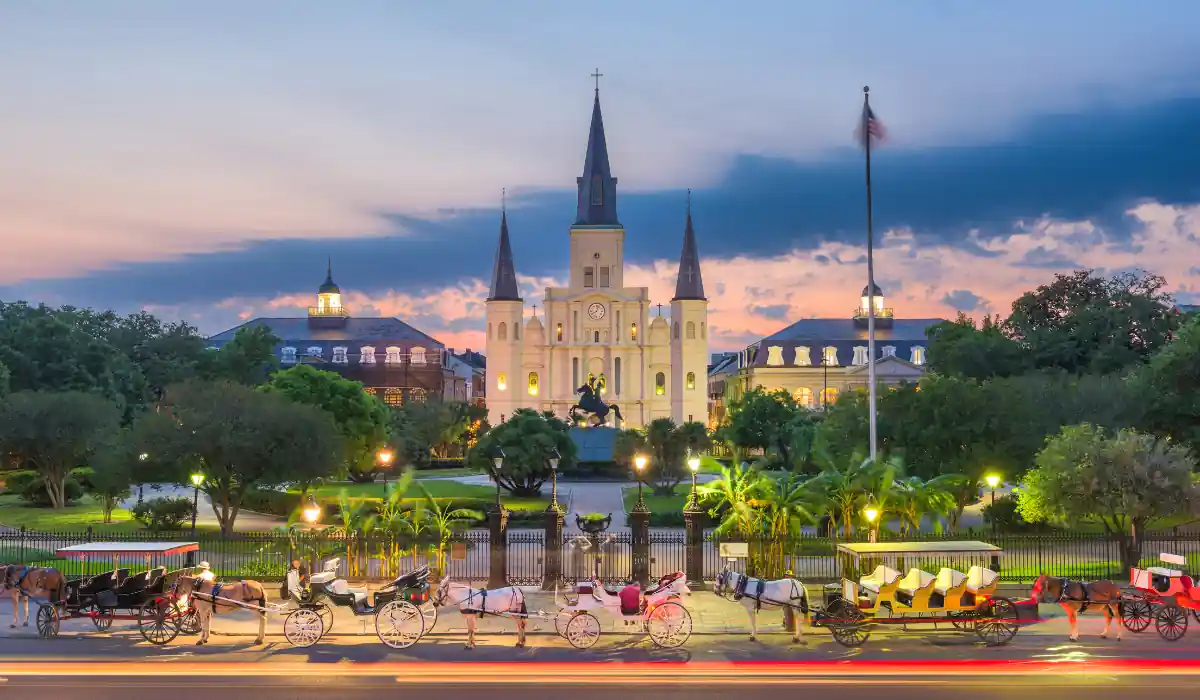 The 15 Best Things to Do in New Orleans for Couples 10 Jackson Square in New Orleans at sunset, with its three spires visible against a colorful sky. Carriages line the street in front, and lush greenery surrounds the area, creating a picturesque and inviting scene.