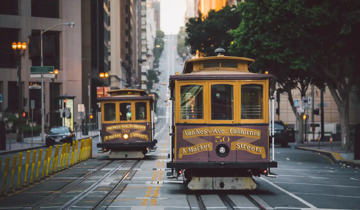 20 Most Romantic Things to do in California for Couples 16 Two vintage San Francisco cable cars travel along a steep, tree-lined street at sunset. The cars are marked with Van Ness Ave. and Market Street, capturing the citys iconic transit system against an urban backdrop.