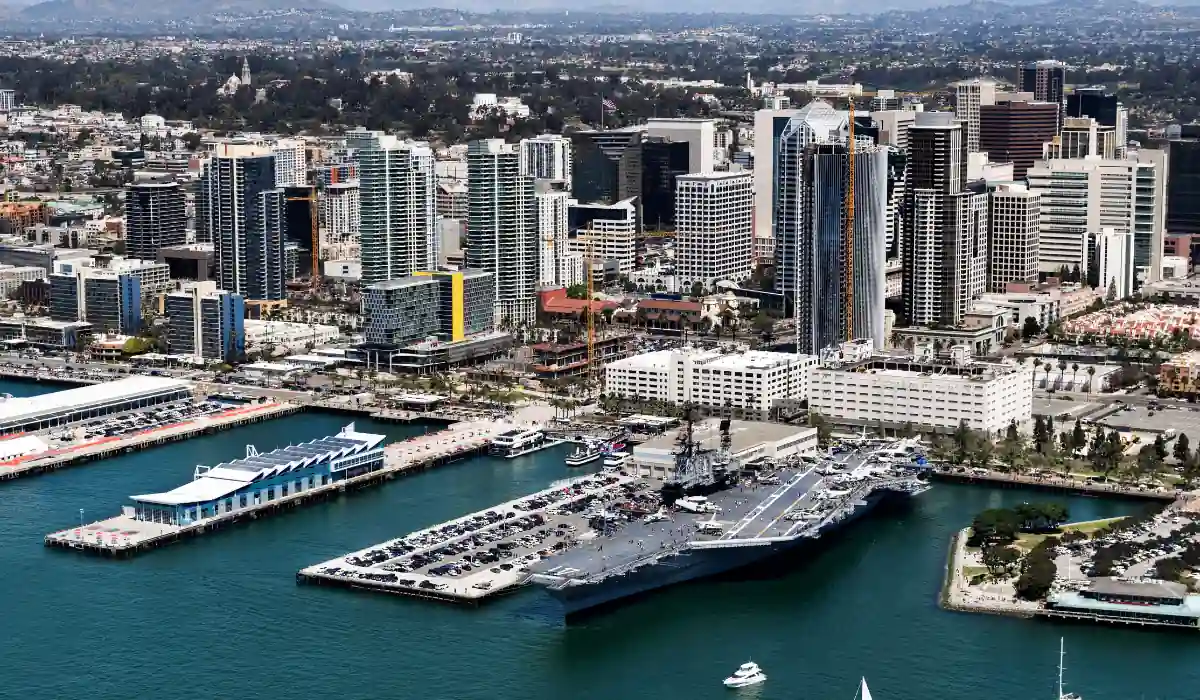 12+ Best Things to do in San Diego for Couples 2025 4 Aerial view of a coastal city with modern skyscrapers and a bay. A large naval ship and a marina are visible in the foreground, while lush hills and sprawling cityscape extend into the background.