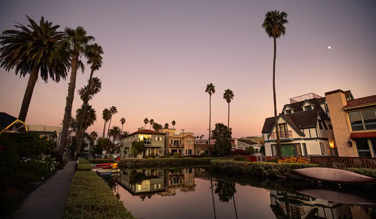 20 Most Romantic Things to do in California for Couples 21 A tranquil canal scene at twilight, with tall palm trees lining the water and charming houses on both sides. The sky is a soft gradient of pink and purple, and a small moon is visible in the sky. Reflections shimmer in the calm water.