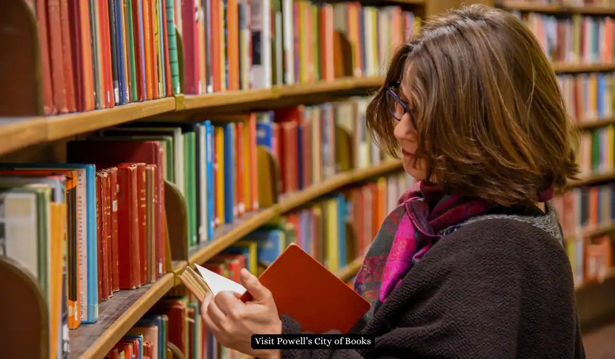 Top 15+ Romantic Things to do in Portland for Couples 10 A person with brown hair and glasses stands reading a book in a library. They are wearing a dark sweater and a purple scarf. Shelves filled with colorful books are on either side.