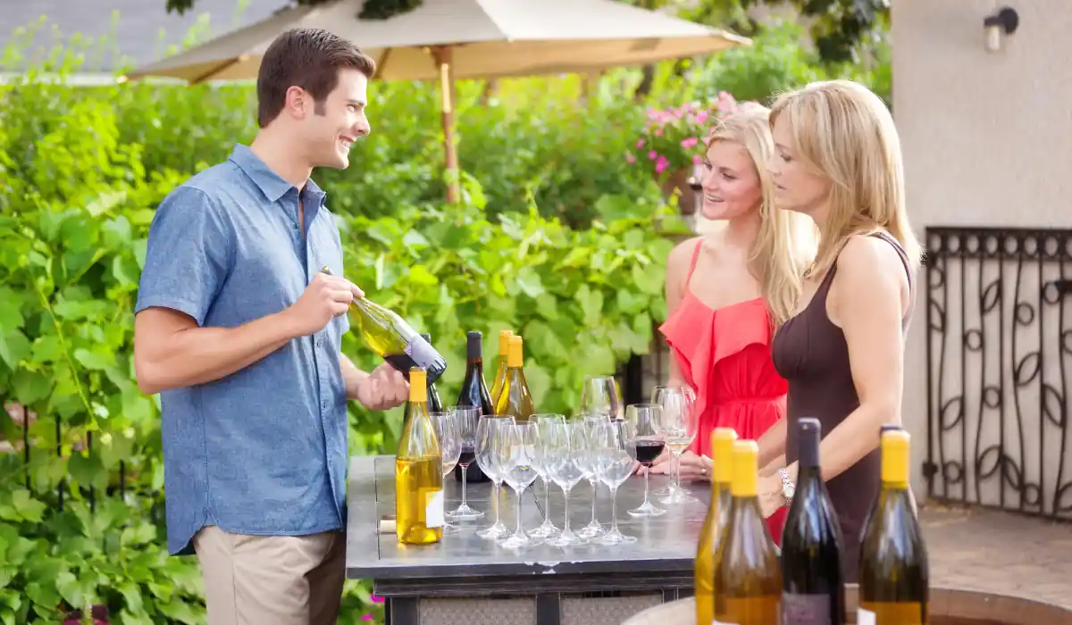 20 Most Romantic Things to do in California for Couples 4 A person pours wine for two women at an outdoor wine tasting event. The table holds several wine bottles and glasses. The setting is a garden with green foliage and a patio umbrella in the background.