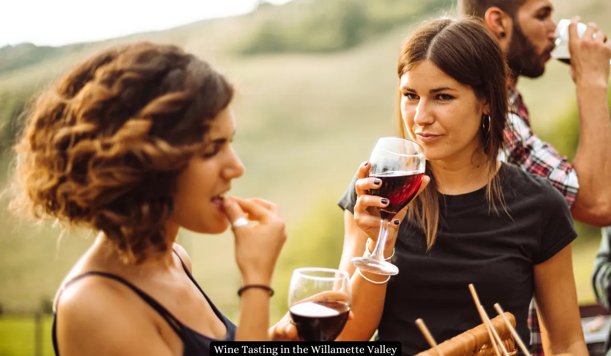 Top 15+ Romantic Things to do in Portland for Couples 5 Two women enjoying a wine tasting event outdoors. One woman holds a glass of red wine while the other samples food. The background features a blurred landscape with greenery. A man is partially visible, sipping from a wine glass.