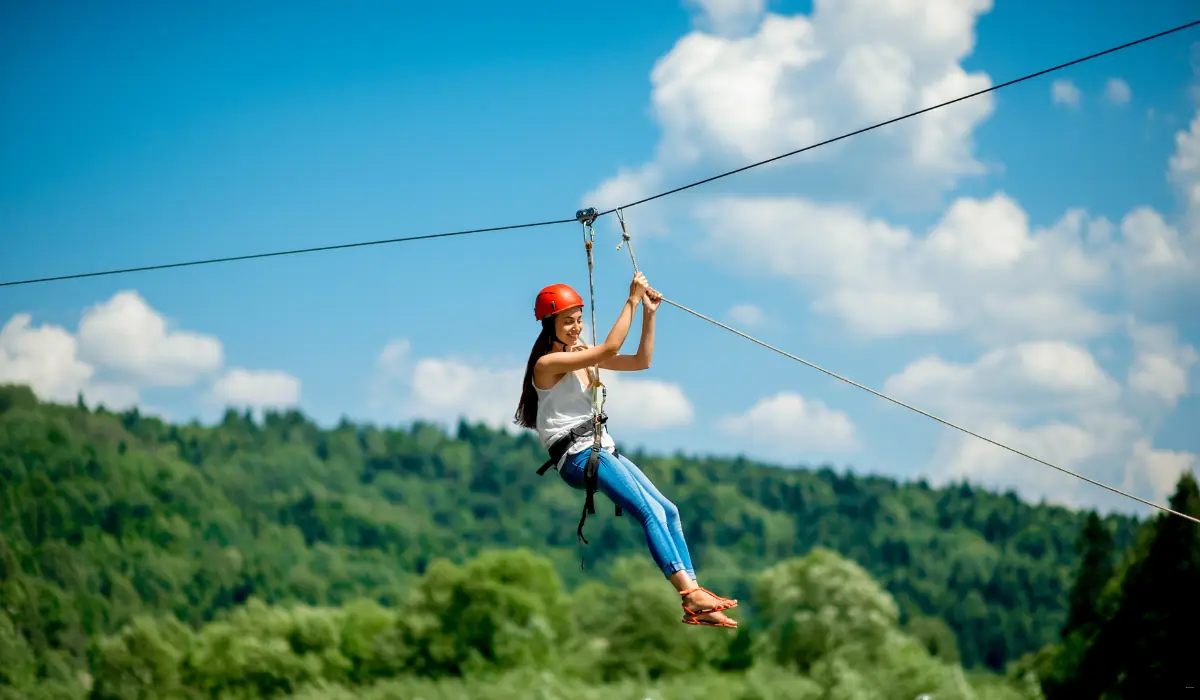 40 amazing Cute Date Ideas in Columbus, Ohio for Couples in 2025 14 A girl wearing a red helmet and harness zip-lining over a lush green forest under a bright blue sky with scattered clouds.