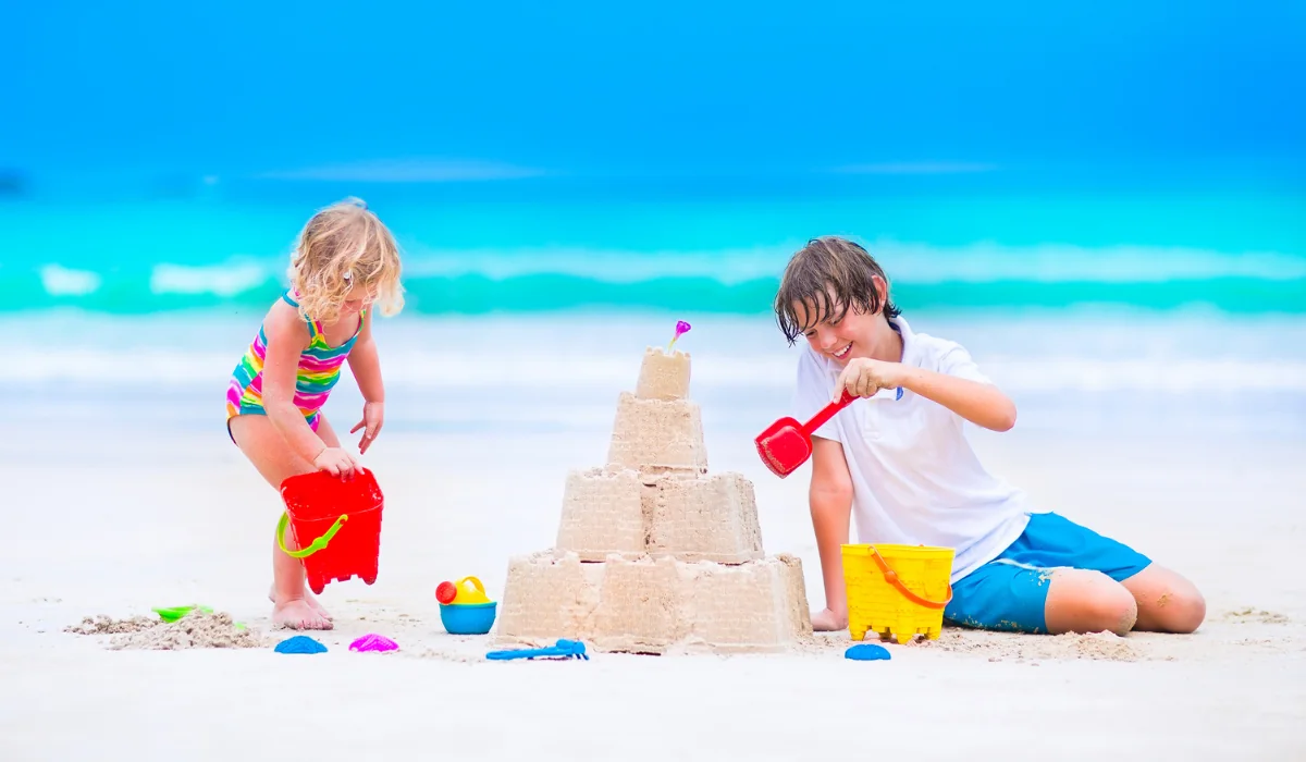 Two children play on a beach. A boy in a white shirt and blue shorts builds a sandcastle with a red shovel, while a younger child in a rainbow-striped swimsuit holds a red bucket. The ocean and sky are vivid blue in the background.
