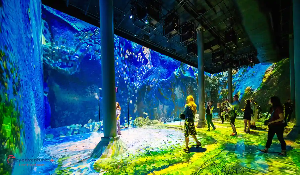 The 10 Best Interactive Museums in NYC You Must Visit  7 People explore an immersive digital art exhibit filled with vibrant blue and green underwater imagery, featuring corals and bubbles. The room has large floor-to-ceiling screens, and the colorful display reflects on the floor and visitors.