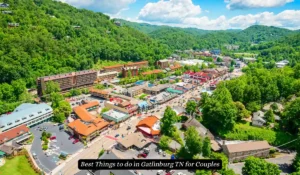 Aerial view of Gatlinburg, TN, featuring a lush green landscape surrounding a vibrant town with red-roofed buildings. The scene includes busy streets and various establishments under a partly cloudy blue sky. "Best Things to do in Gatlinburg TN for Couples"