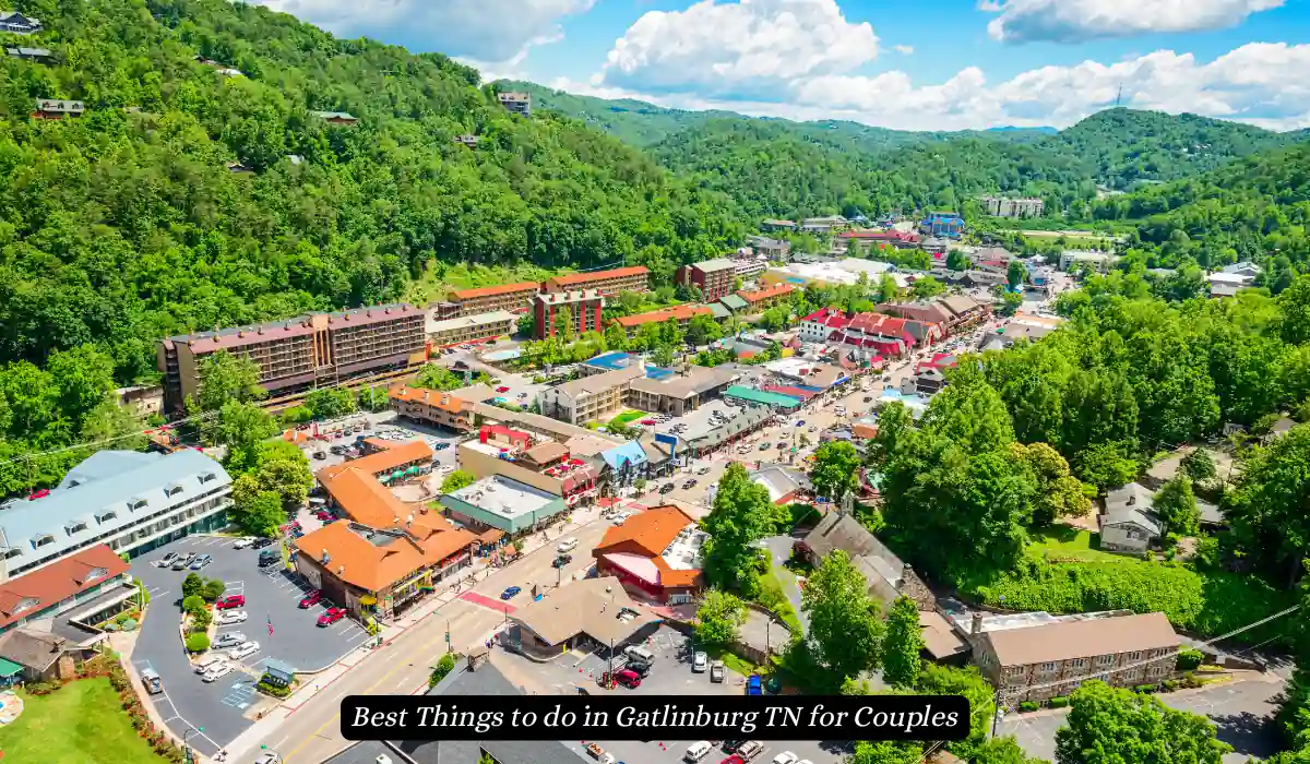 Aerial view of Gatlinburg, TN, featuring a lush green landscape surrounding a vibrant town with red-roofed buildings. The scene includes busy streets and various establishments under a partly cloudy blue sky. "Best Things to do in Gatlinburg TN for Couples"
