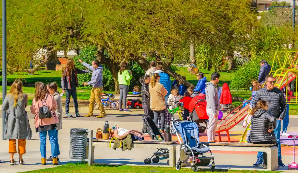A Brackenridge park scene with adults and children enjoying a sunny day. People are gathered, some interacting while others are near a stroller. Trees and a playground area provide a lively backdrop under a clear blue sky.
