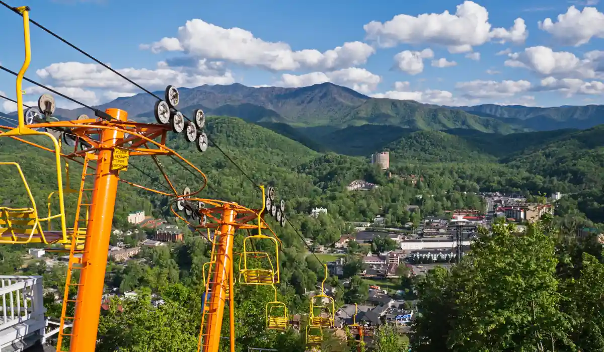 Top 15 Best Things to do in Gatlinburg TN for Couples 3 A scenic view of a mountainous landscape with a bright yellow chairlift in the foreground. Below, a town nestled in lush green forests is visible. The sky is dotted with fluffy clouds and mountains stretch across the horizon.
