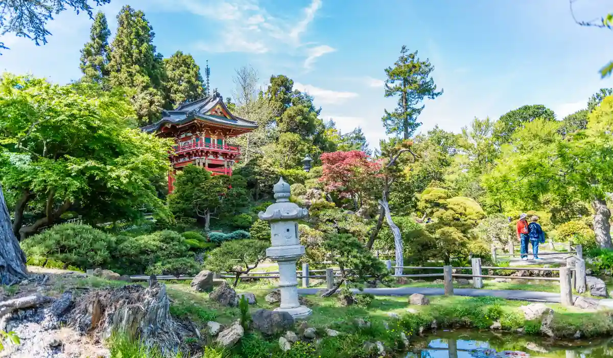 A tranquil Japanese garden scene with a red pagoda, lush greenery, and a stone lantern by a pond. Two people walk along a gravel path under a bright blue sky.

