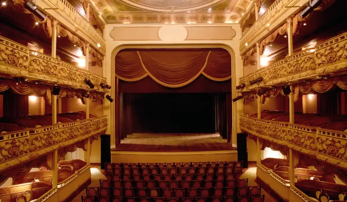 Interior of an ornate theater featuring red velvet curtains and golden ornate balconies. Rows of empty seats face the stage, with warm lighting creating an elegant atmosphere.
